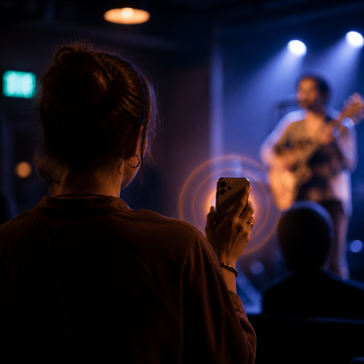 Someone holding up a phone at a small live show while a guitarist plays under stage light.